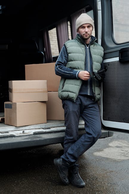A man dressed in a green padded vest, navy blue jacket, grey beanie, and dark trousers leans casually against the open side of a grey van during a home relocation process. Behind him, inside the van, 