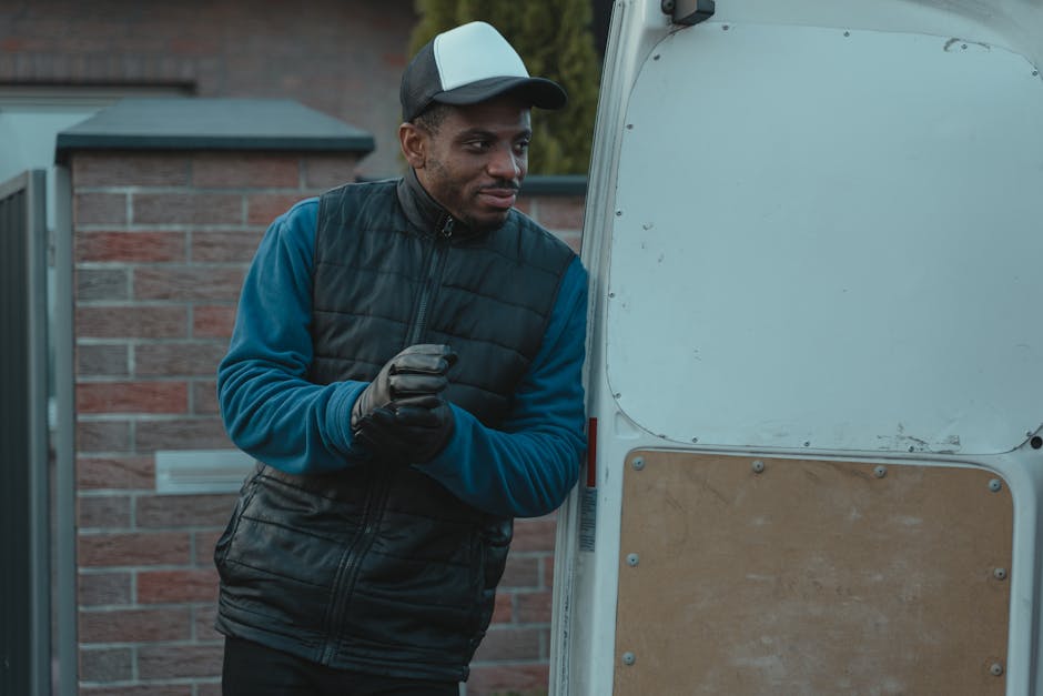 A man dressed in a green padded vest, navy blue jacket, grey beanie, and dark trousers leans casually against the open side of a grey van during a home relocation process. Behind him, inside the van, 