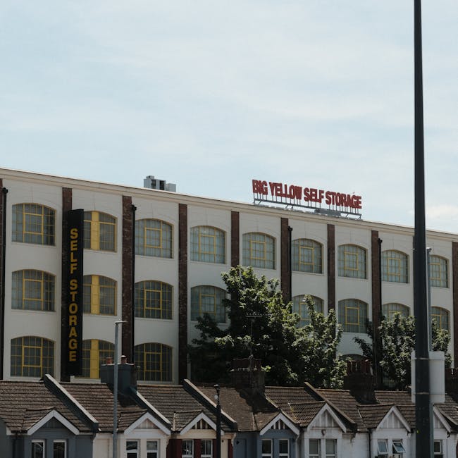 Exterior view of a multi-storey white building with large arched windows framed in yellow, featuring signs for 'SELF STORAGE' and 'BIG YELLOW SELF STORAGE' on the facade. In front of the building, there are smaller houses with tiled roofs, and a few trees partially obscure the lower portion of the structure. A tall street lamp is visible to the right. The scene is captured during daylight with a partly cloudy sky, illustrating a commercial storage facility typical for relocation and moving services provided by Man with Van Swiss Cottage, supporting home relocation and furniture transport within the area.